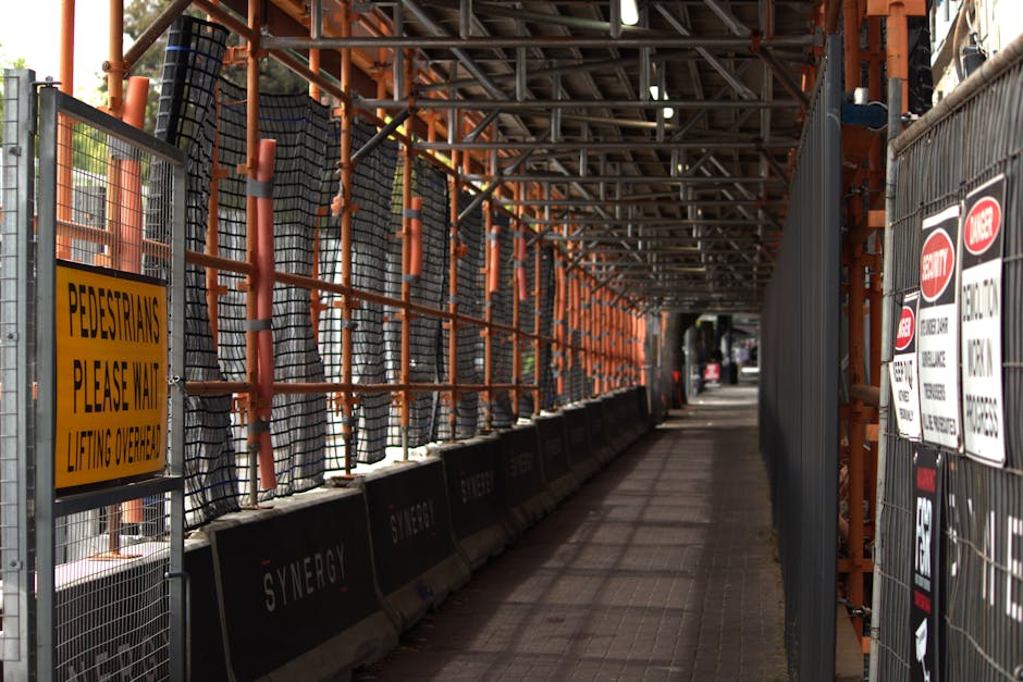 A long, narrow pedestrian pathway adjacent to a construction site, enclosed by metal fencing and orange safety scaffolding. The fencing features black mesh panels and various warning and informational